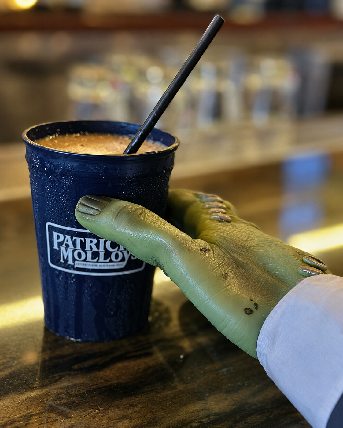 A green monster hand reaching for a navy Patrick Molloy's cup holding a frozen Irish coffee at the bar.