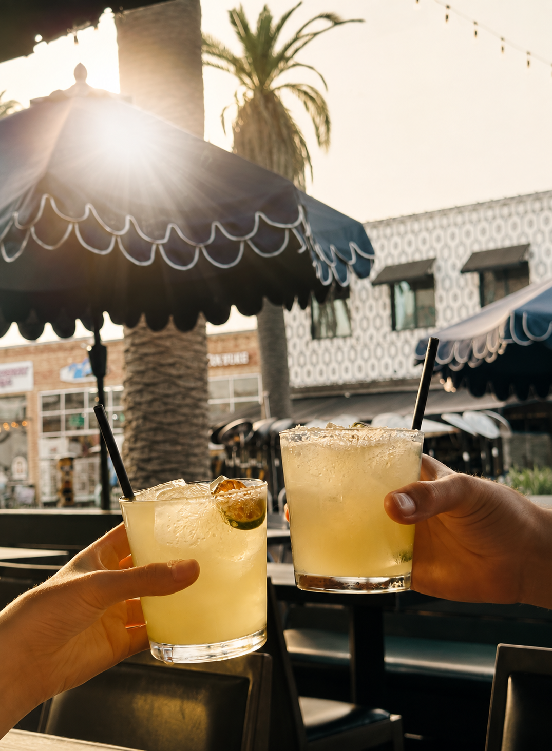 Two hands clinking margaritas over ice under a scalloped patio umbrella at golden hour.