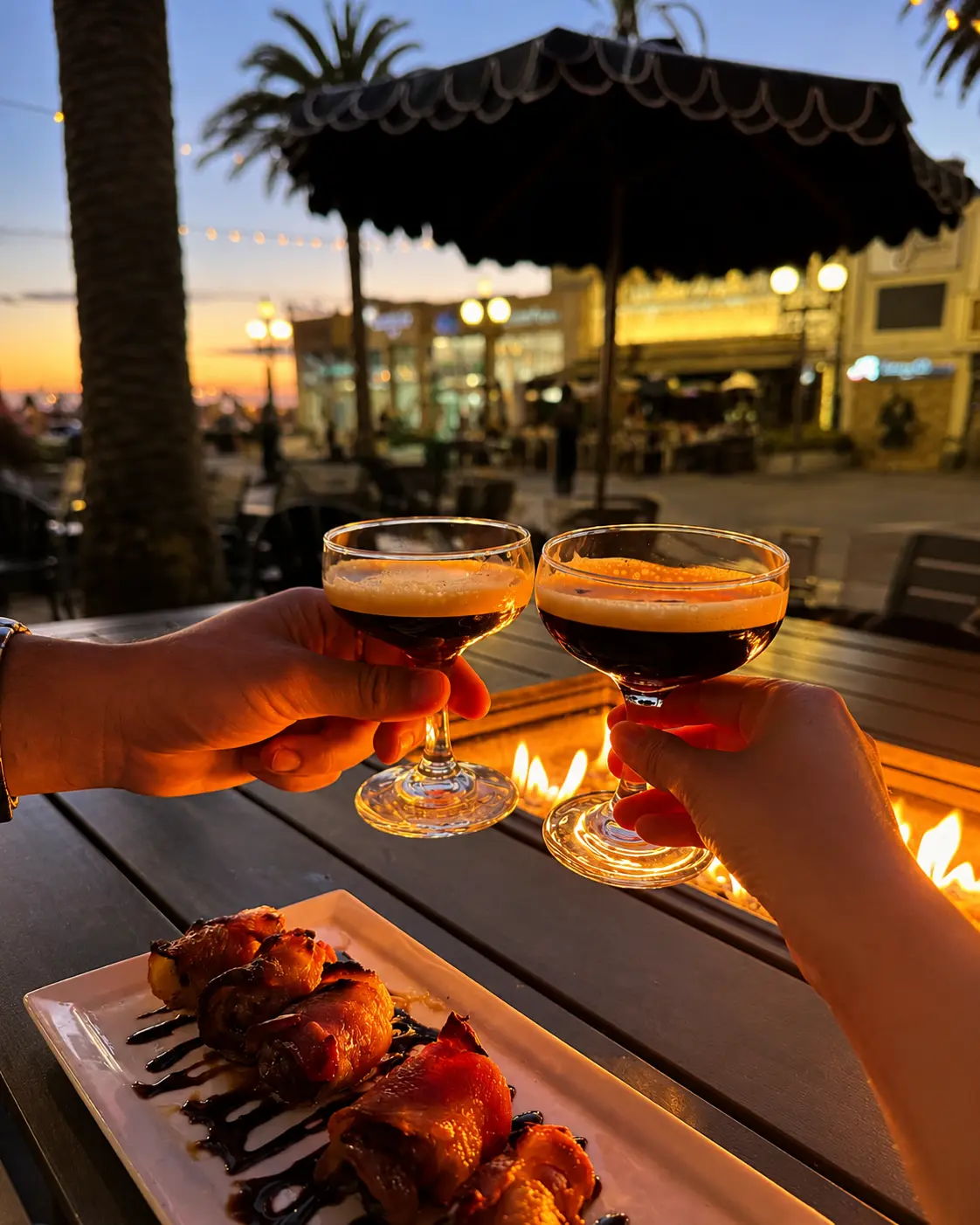 Two hands toasting espresso martinis over a lit fire table at sunset on the Patrick Molloy's patio.