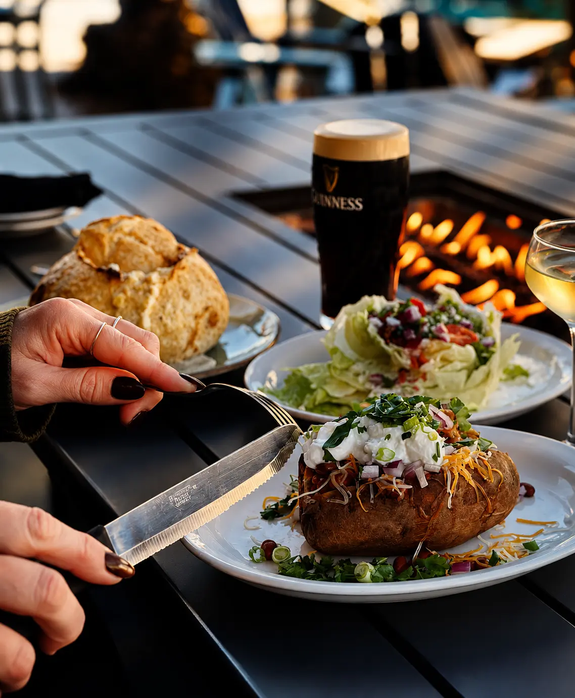 A loaded baked potato with chili, cheddar, sour cream and scallions on a patio table beside a pint of Guinness and a fire pit at sunset.
