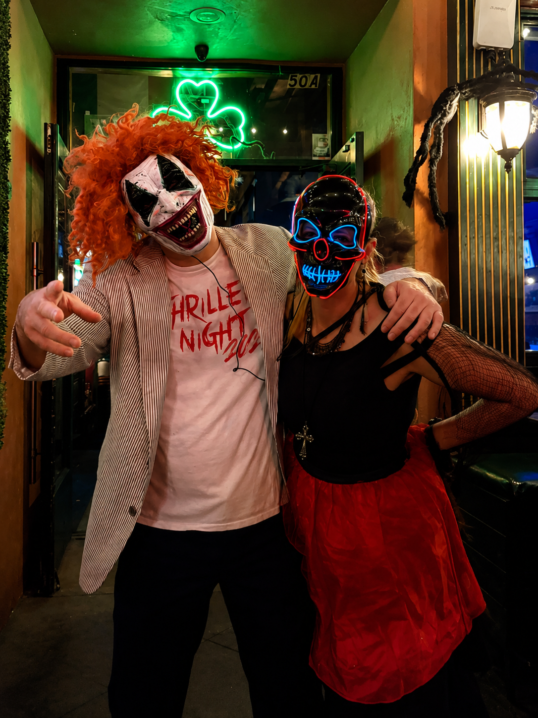 Two guests in a clown mask and a glowing LED skull mask posing under the green neon shamrock at the entrance.