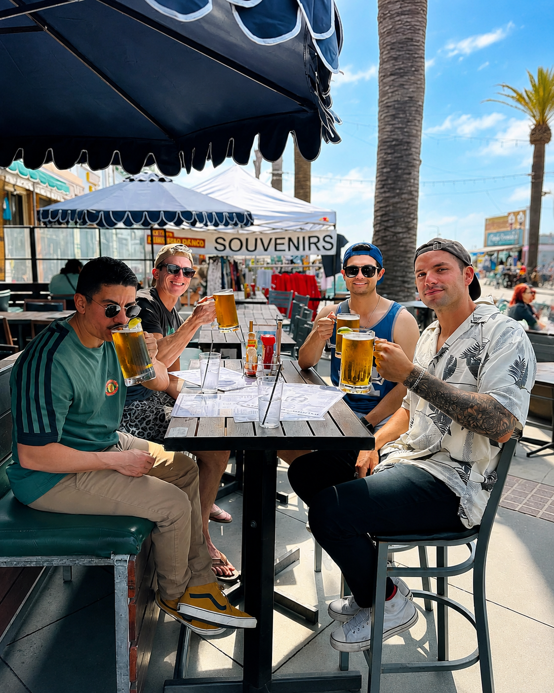 Four friends at an outdoor patio table on a sunny day raising tall glass beer steins together.