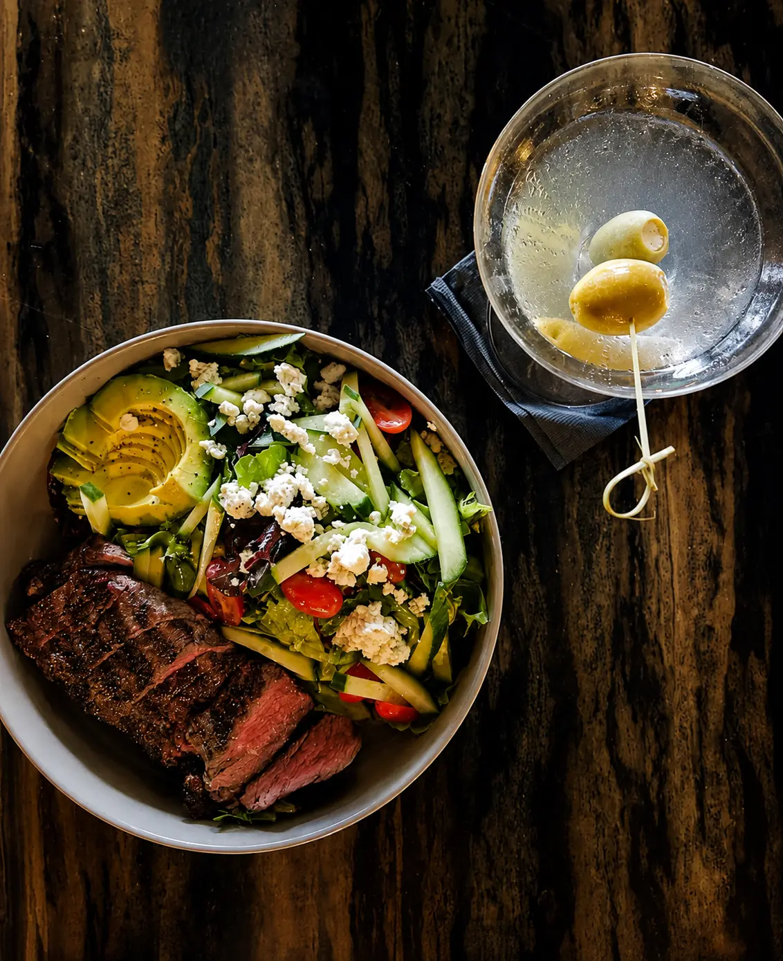 Overhead view of a bowl of greens with sliced steak, avocado, cucumber, cherry tomatoes and bleu cheese, beside a martini with olives.