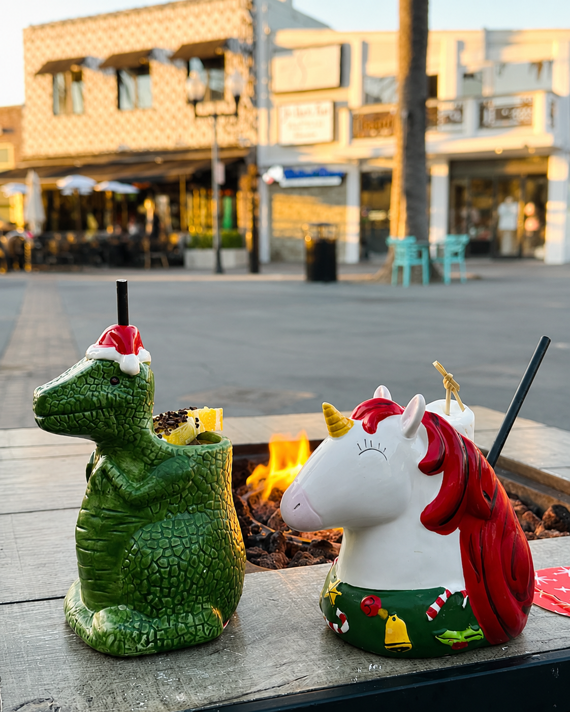 Two collectible holiday tiki mugs, a green dinosaur and a red-maned unicorn, on a fire table at sunset on Pier Ave.