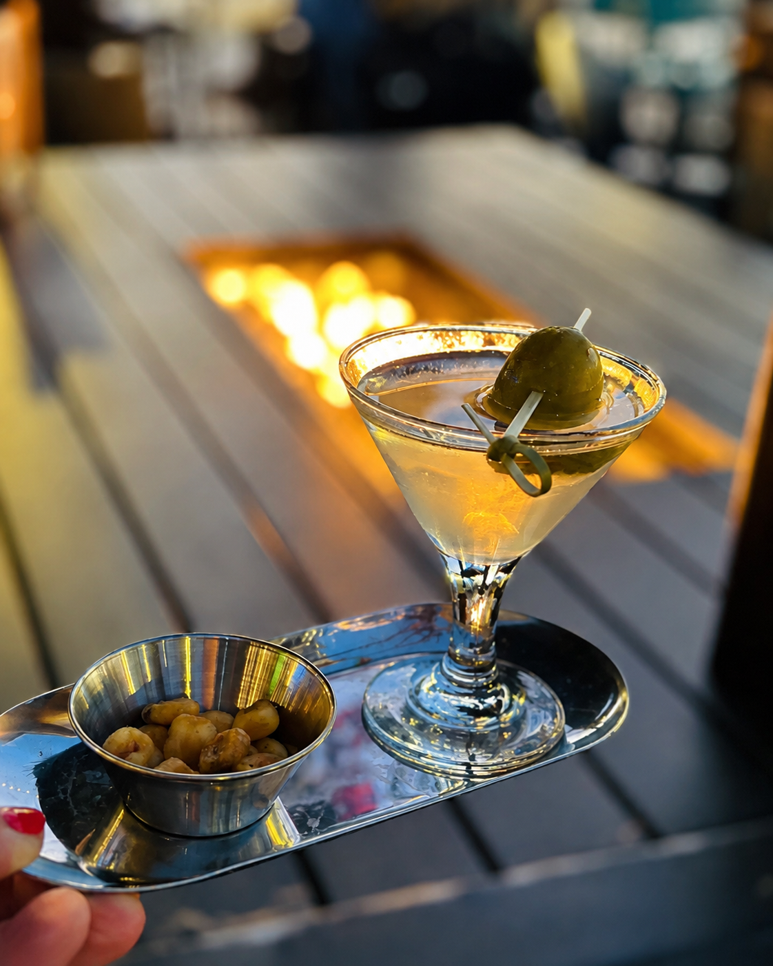 A martini with a blue-cheese-stuffed olive on a silver tray with a cup of marinated olives, carried past a lit fire table.