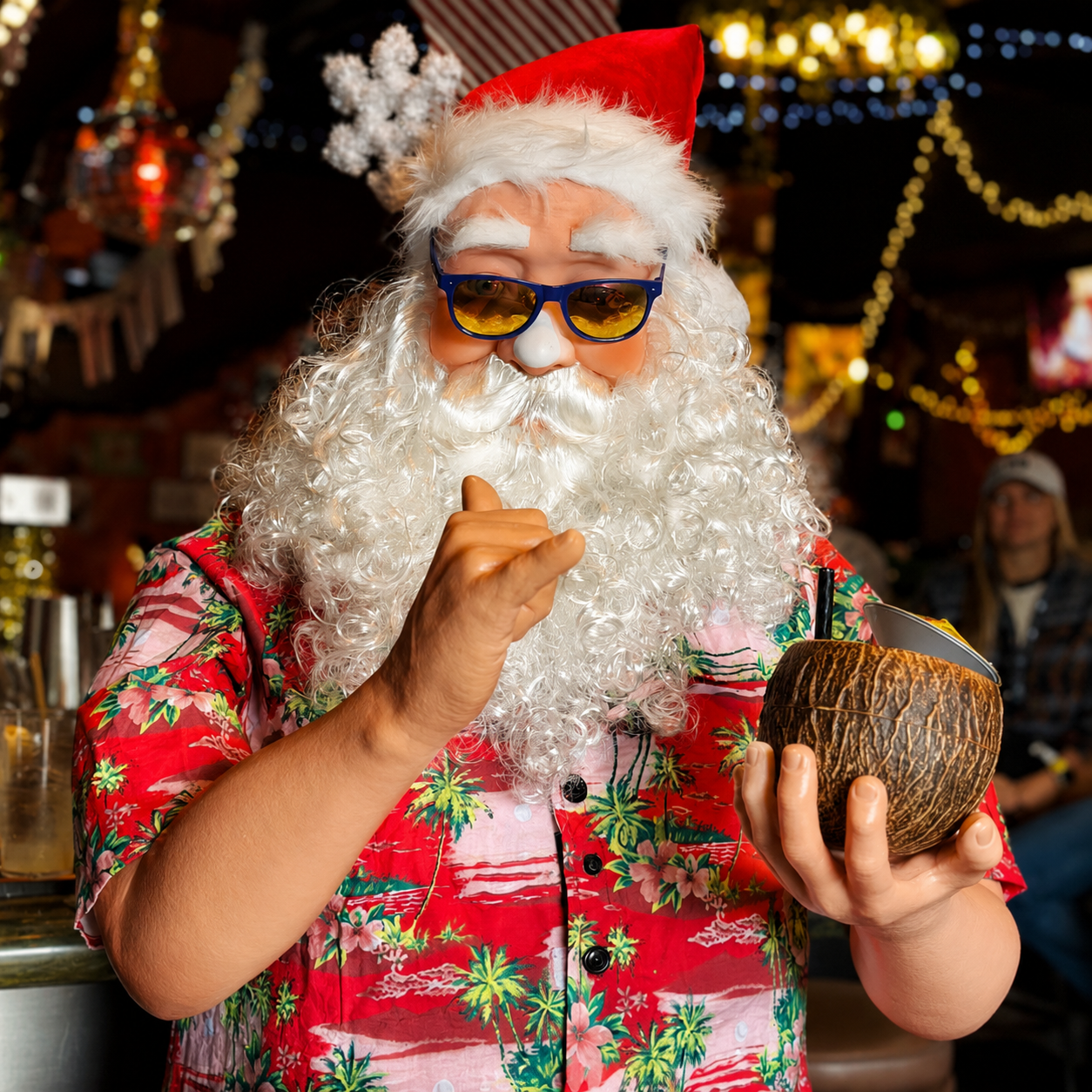 A beachy Santa in a Hawaiian shirt and sunglasses holding a coconut cocktail under holiday lights.